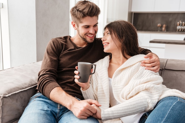 Cheerful lovely young couple sitting and drinking coffee on sofa at home