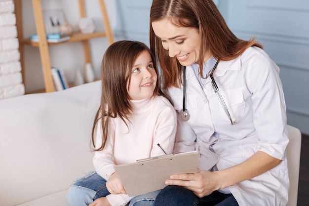 Cheerful loving mother making prescriptions for her little daughter while sitting together on the couch