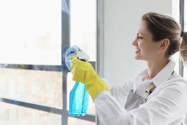 Cheerful maid sprays detergent on window glass in hotel room woman in rubber gloves and apron