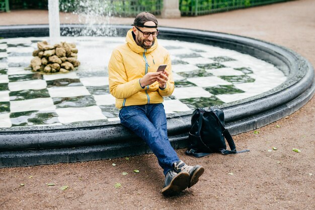 Cheerful man in cap eyeglasses and yellow anorak sitting near fountain chatting with friends using mobile phone Tired man after walk in big city having rest near fountain watching video online