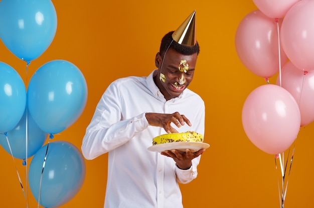 Cheerful man in cap with smeared face tasting birthday cake. Smiling male person got a surprise, event celebration, balloons decoration