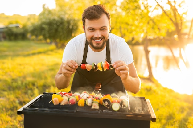 Cheerful man cooking skewers on lake coast