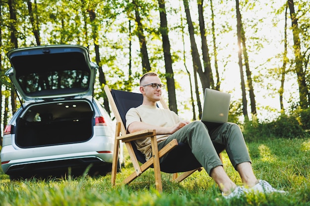 A cheerful man in glasses sits on a chair in nature and works on vacation work online on a portable laptop Freelance work