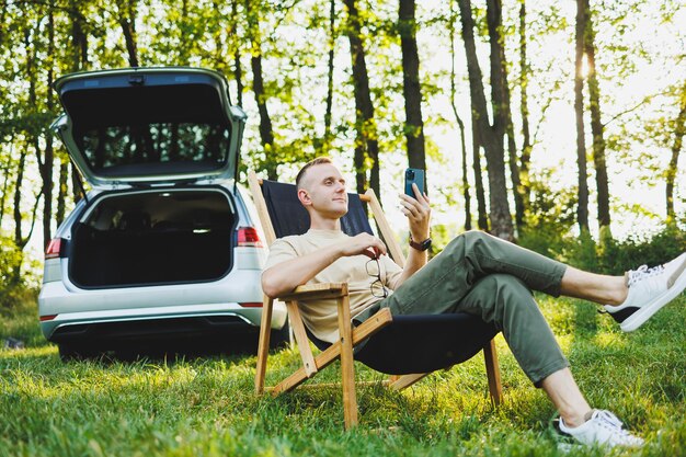 A cheerful man in glasses sits on a chair in nature and works on vacation work online on a portable laptop Freelance work