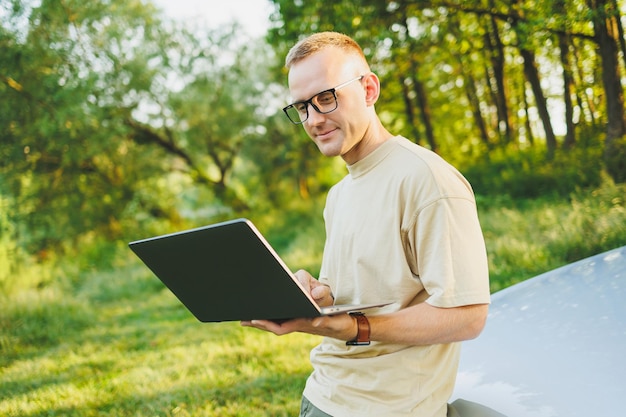 A cheerful man in glasses sits on the hood of his car in nature and works on a laptop Freelance work remote work during vacation