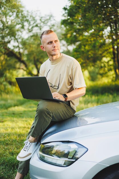 A cheerful man in glasses sits on the hood of his car in nature and works on a laptop Freelance work remote work during vacation