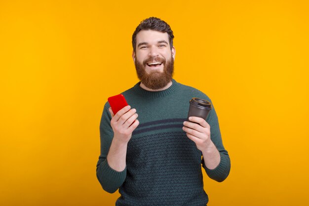 Cheerful man is holding a paper cup and his phone on yellow background.