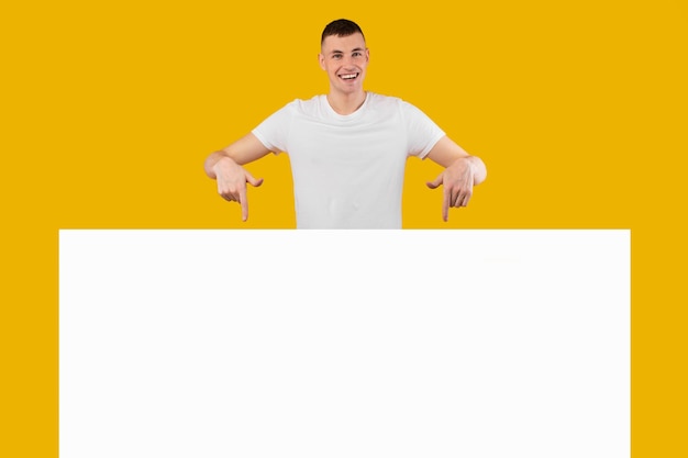 Cheerful man pointing fingers at empty paper board yellow background