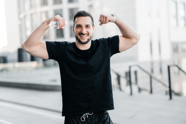 Cheerful man showing his muscles. Hard workout concept. Fit man showing biceps and smiling. Gray morning city on background. Handsome man excercised a lot to get a fit body.