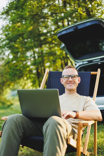 A cheerful man sits on a chair in nature and works online on a laptop A man travels and works remotely on a laptop computer Office work in nature