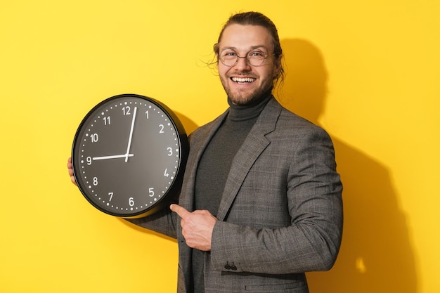 Cheerful man wearing glasses holding big clock on yellow background