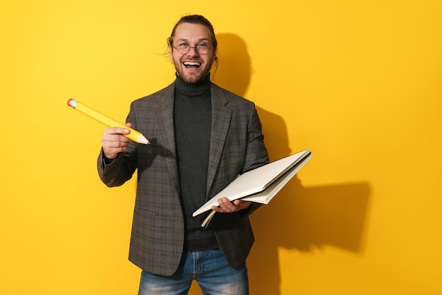 Cheerful man wearing glasses holding big pencil and notebook on yellow background