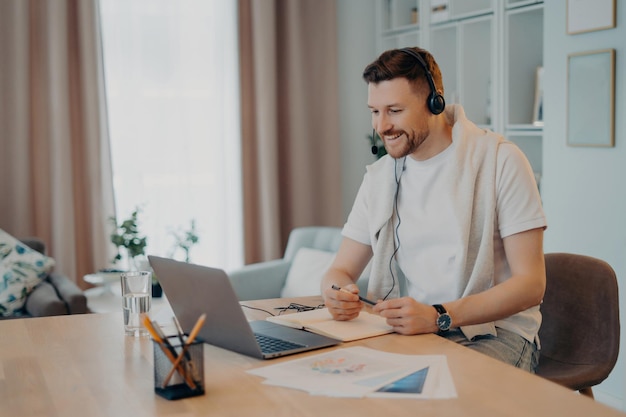 Cheerful man with headphones working on laptop at home taking notes in a bright cozy room