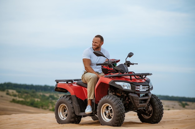 Cheerful man with helmet in hand poses on atv, downhill riding in desert sands. Male person on quad bike, sandy race, dune safari in hot sunny day, 4x4 extreme adventure, quad-biking