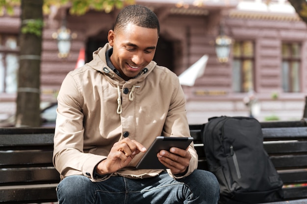 Cheerful man with tablet sitting on wooden bench near backpack and chatting