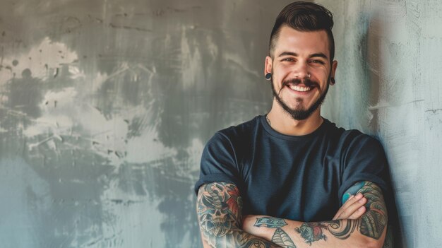 A cheerful man with tattoos leans against a rustic wall exuding confidence and warmth with his arms crossed and a bright smile