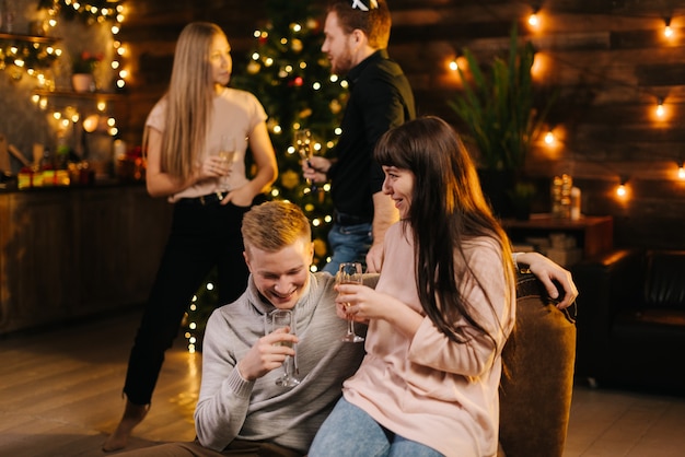 Cheerful man and woman enjoying conversation in festively decorated house holding glasses of champagne in hand. Young couple talking on New Year Eve against backdrop of festive Christmas tree.