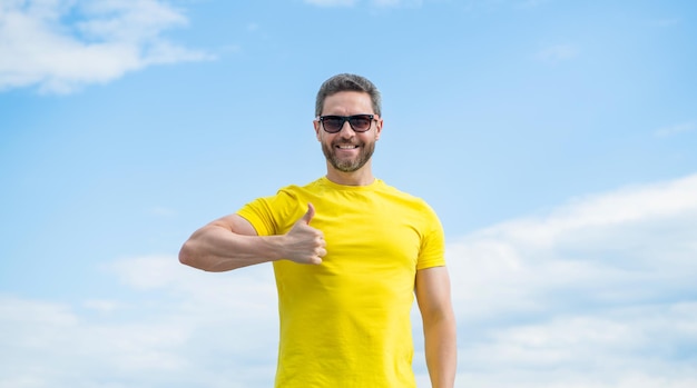 Cheerful man in yellow shirt and sunglasses outdoor on sky background thumb up