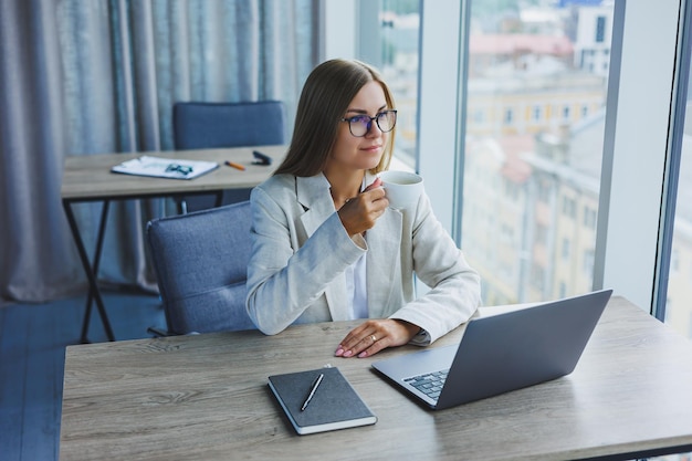 Cheerful manager woman in glasses with laptop smiling at camera during coffee break in office interior happy european woman with caffeine drink posing