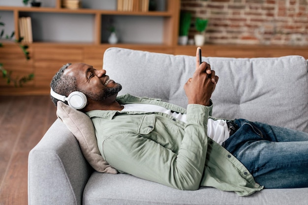 Cheerful mature african american male in casual in wireless headphones enjoys favorite music with smartphone