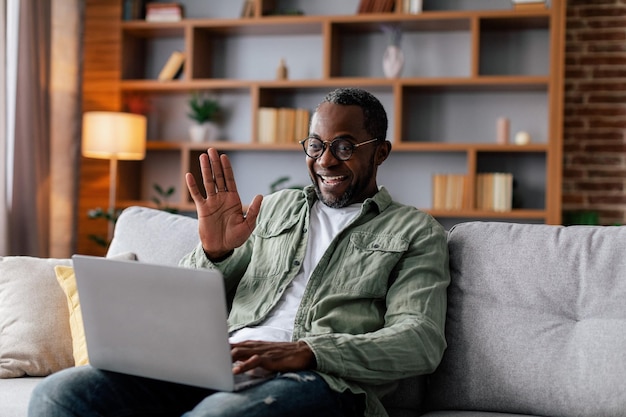 Cheerful mature african american male in glasses and casual waving hand and looking at computer