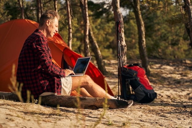 Cheerful mature man is sitting near tent in nature and typing on laptop after nordic walking