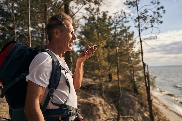 Cheerful mature man with tourist backpack is going walking in forest near sea and communicating on smartphone