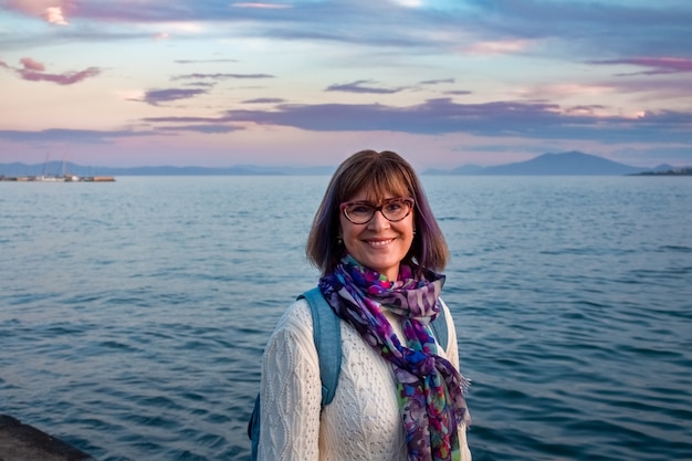 Cheerful mature woman with spectacles wearing white sweater and colorful scarf smiling near the sea behind her in Greece. Happiness and vacation concept.