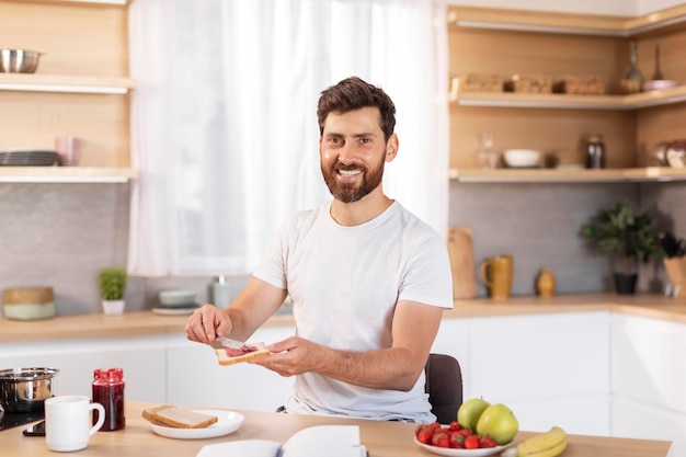 Cheerful middle aged caucasian male with beard in white tshirt makes sandwich with fruits for breakfast