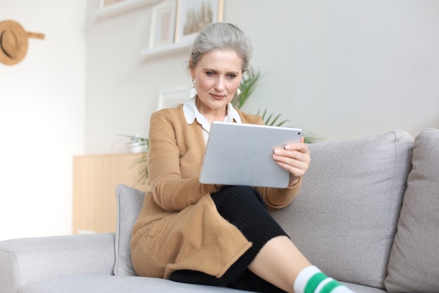 Cheerful middle aged woman sitting on sofa, using computer tablet apps, looking at screen,reading good news in social network, shopping or chatting online.