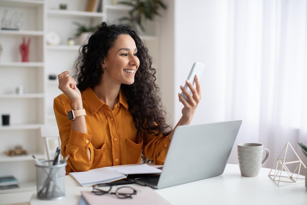 Cheerful middle eastern businesswoman using cellphone sitting at workplace