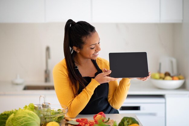 Cheerful millennial african american female in apron show tablet with empty screen