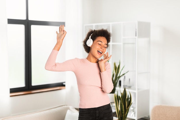 Cheerful millennial african american woman in wireless headphones sings song enjoys favorite music dancing