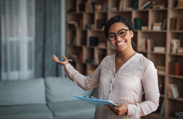 Cheerful millennial black female doctor psychologist in glasses with tablet invites client to modern