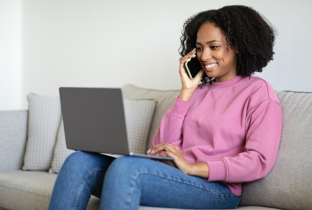 Cheerful millennial black woman typing on computer reads message calls by phone sits on sofa