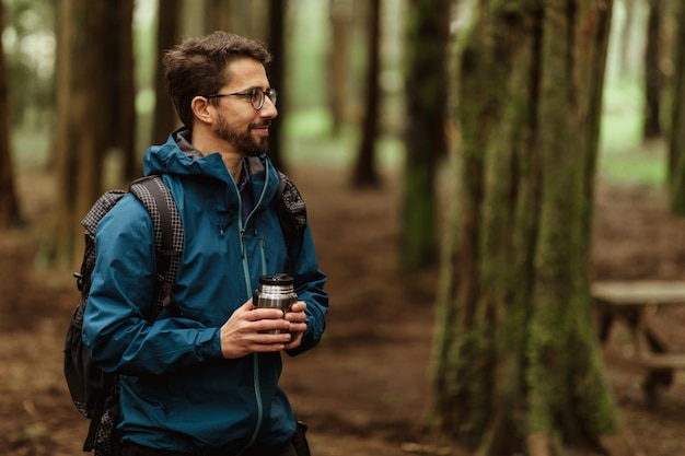 Cheerful millennial caucasian man in glasses jacket with backpack and cup of hot drink resting in cold forest