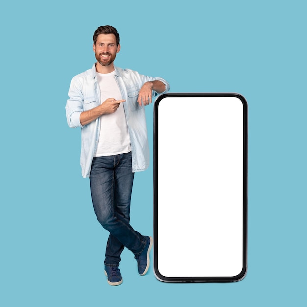 Cheerful millennial caucasian man point finger at huge smartphone with blank screen