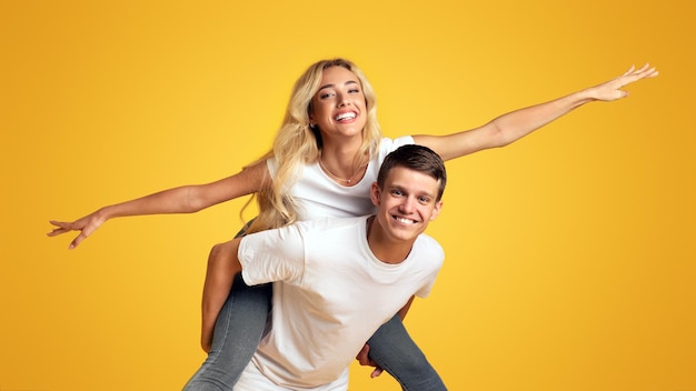 Cheerful millennial couple having fun fooling together over yellow background girl riding her