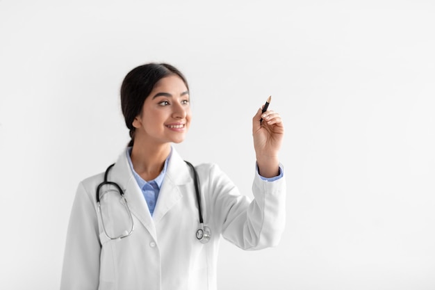 Cheerful millennial hindu lady therapist in coat writes with pen on empty space