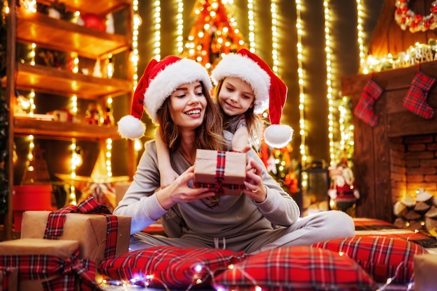 Cheerful mom and her cute daughter girl exchanging gifts. Parent and little children having fun near tree indoors. Loving family with presents in christmas room.