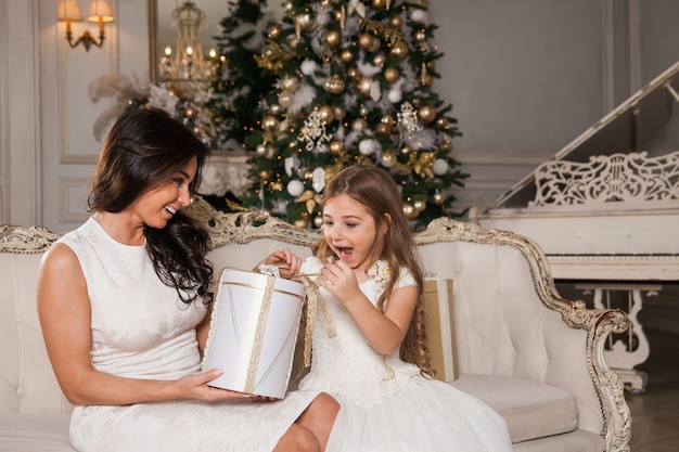 Cheerful mom and her cute daughter girl exchanging gifts in white classic interior against the background of a piano and a decorated Christmas tree. 