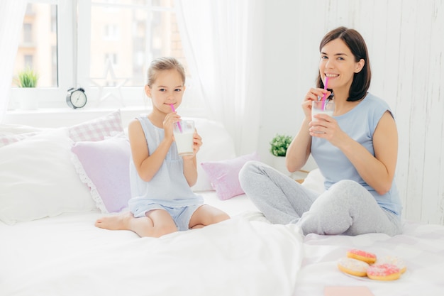 Cheerful mother and daughter dressed in pyjamas, have breakfast in morning