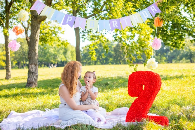 Cheerful mother and daughter having fun on child birthday on blanket with paper decorations in the park