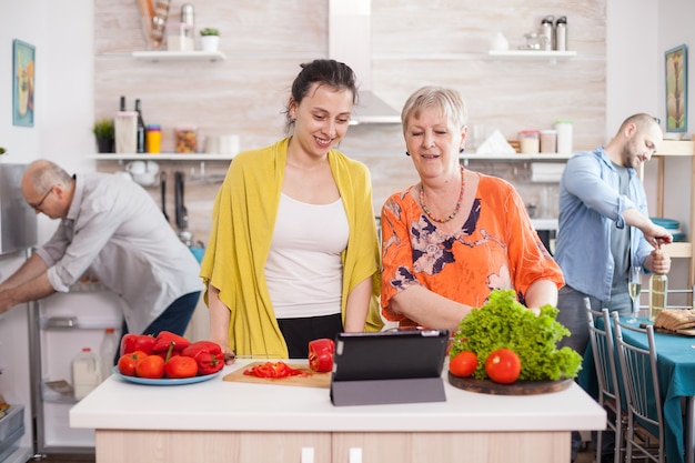 Cheerful mother and daughter using tablet to cooking tasty salad following online recipe in kitchen. Husband holding wine bottle. Father looking inside refrigerator