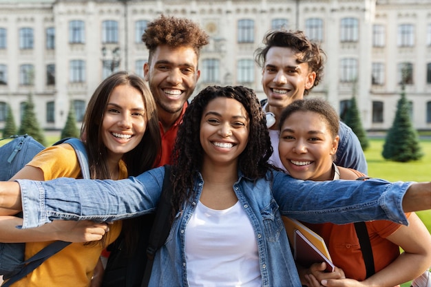 Cheerful multicultural students making selfie near university building posing together outdoors