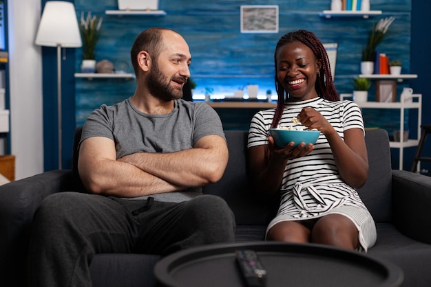 Cheerful multiethnic couple talking and having a snack in the living room. People sitting on sofa while telling jokes, laughing together and bonding on the couch while eating popcorn at home