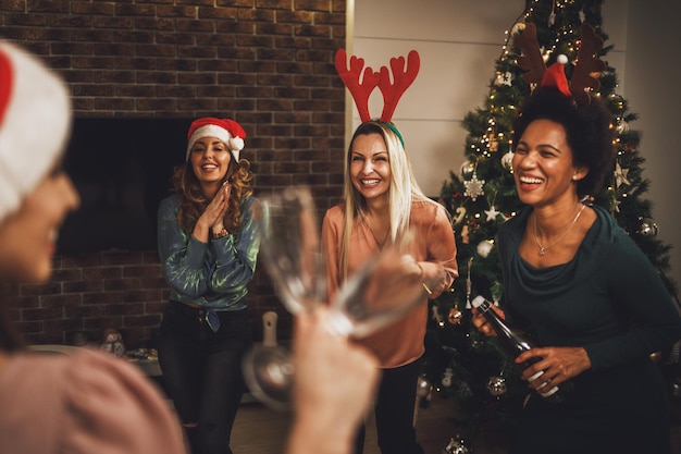 Cheerful multiethnic female friends having fun and making a toast with champagne while celebrating New year or Christmas at home together.