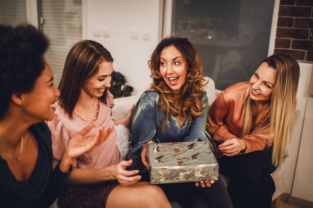 Cheerful multiethnic girls giving presents to each other while gathering at home to celebrate Christmas or New Year.