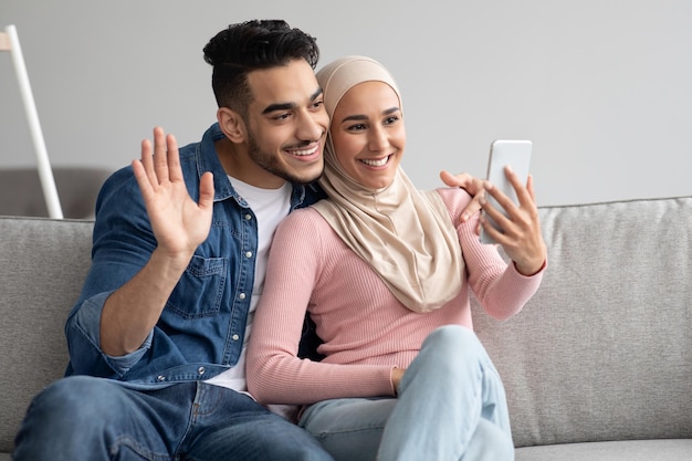 Cheerful muslim family husband and wife having video conference with friends or relatives from home, waving at smartphone screen, sitting on couch in living room, copy space