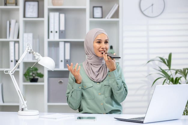 A cheerful muslim woman wearing a hijab speaks into her phone recording an audio message at her